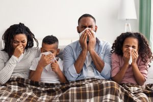 Mother, son, father and daughter sitting under a blanket blowing their noses with tissues.
