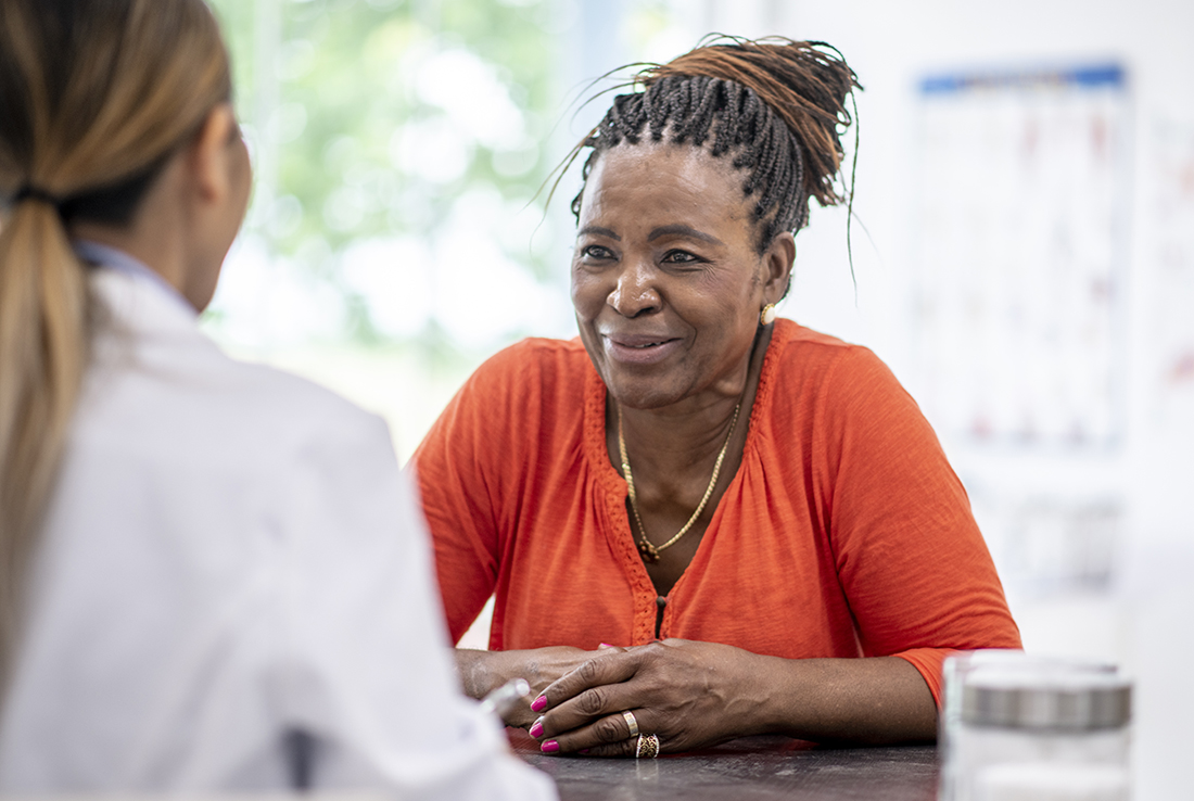 African American woman in an orange top speaks with a female physician at a desk