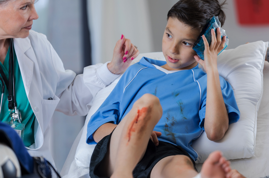 A little boy in a blue hospital gown with a bloody knee is attended to by a doctor